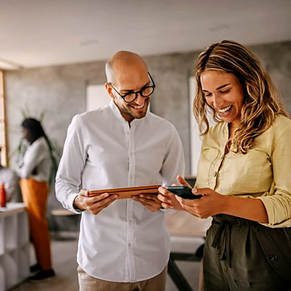 Two colleagues smiling while looking at a phone and tablet together in an office