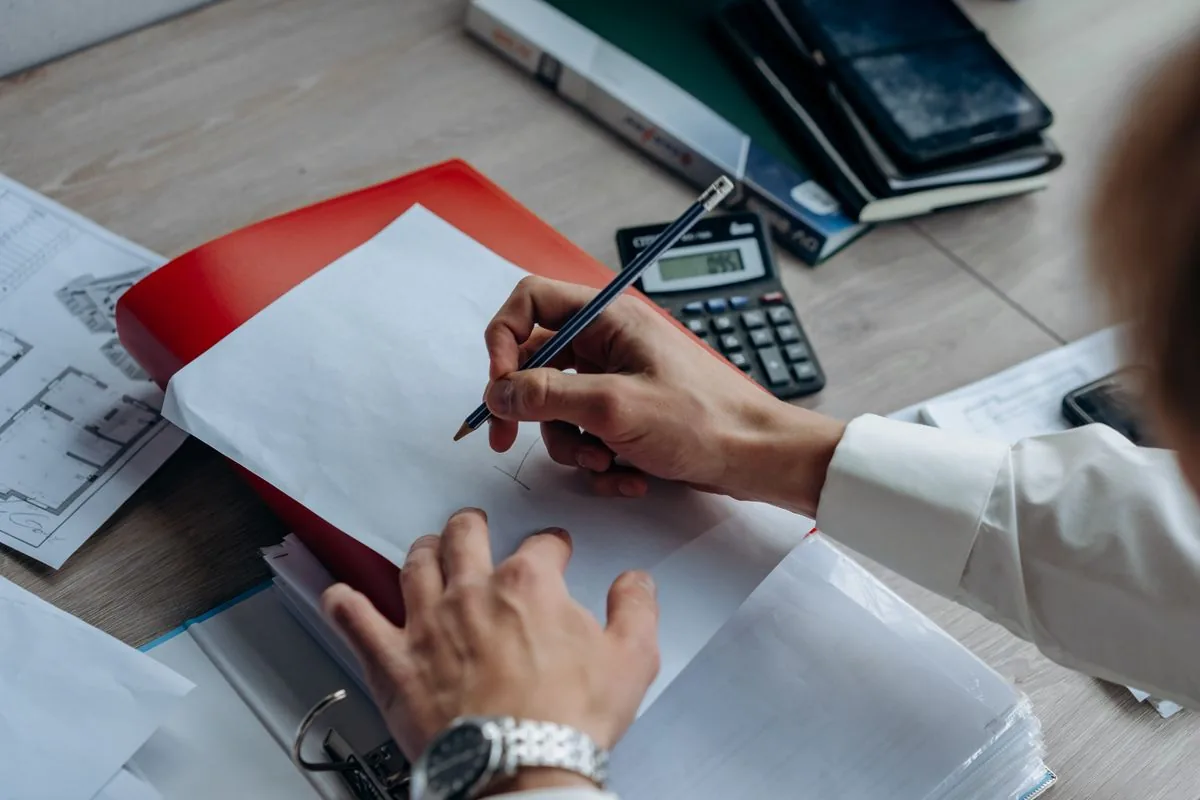 Hands writing in a notebook with a calculator and job folder