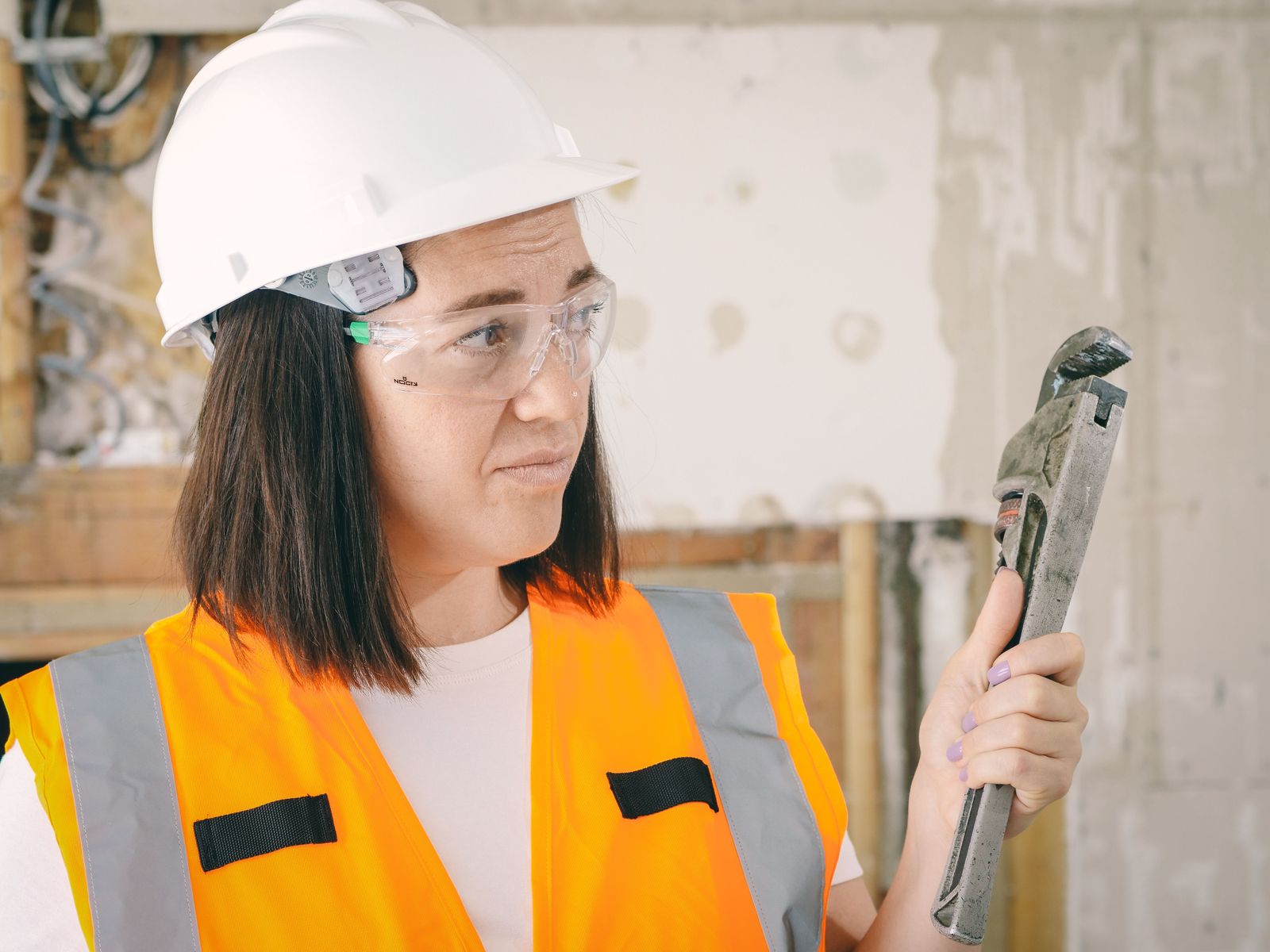 Female trade worker in a hard hat, safety glasses, and hi-vis vest holding a plumber's wrench